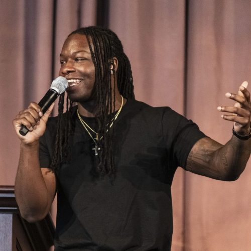 A man with long dreadlocks, wearing a black t-shirt and gold chains, speaks enthusiastically into a microphone about educational enrichment while gesturing with his hand in front of a brown curtain.