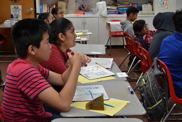 Students sit at desks in a classroom, focusing on something out of view. Some have math papers, whiteboards with graphs, and geometric shapes—tools for educational enrichment. Books and supplies are visible in the background.