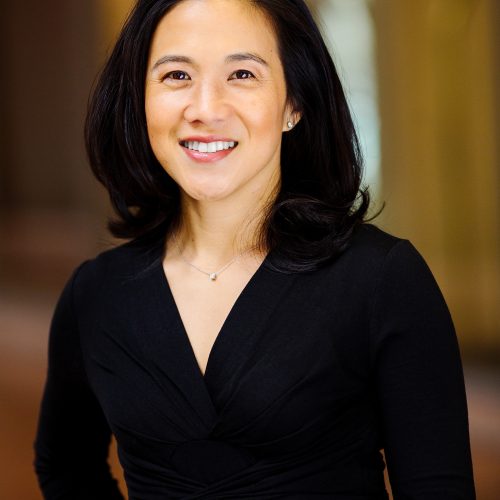 A woman with shoulder-length black hair, wearing a black wrap dress and a small necklace, is smiling and standing in a softly lit indoor setting, embodying educational empowerment against a blurred background.