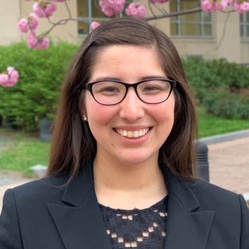 A woman with long brown hair and glasses smiles while standing outdoors in front of blooming pink flowers, embodying educational empowerment in her black blazer and black lace top.