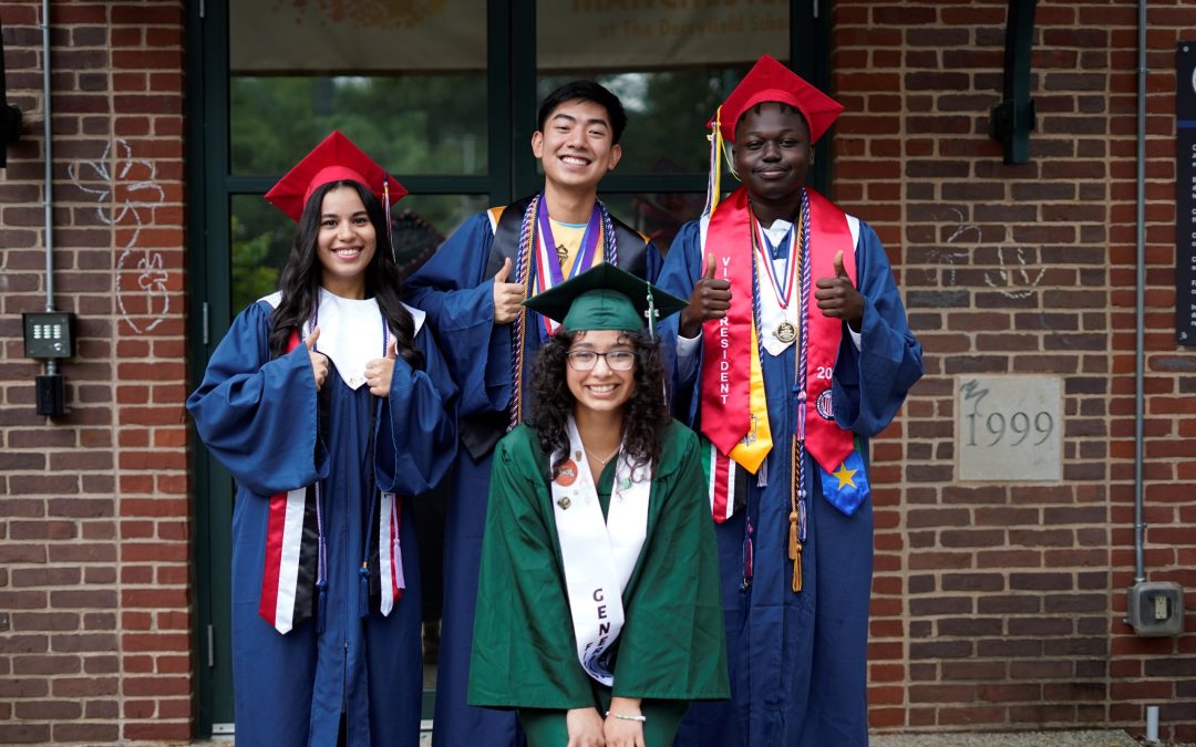 Four smiling graduates in caps and gowns pose in front of a brick building, celebrating educational empowerment. Three wear blue and red behind a fourth in green, who crouches holding a diploma folder while two give thumbs up.