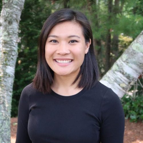 A woman with straight, dark shoulder-length hair wearing a black top smiles while standing outdoors in front of trees and greenery, radiating a sense of educational empowerment.