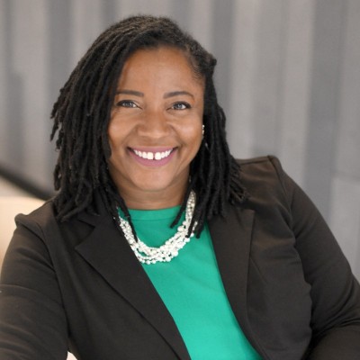 A woman with shoulder-length dreadlocks, wearing a black blazer, green top, and layered pearl necklace, smiles at the camera in front of a blurred gray background, embodying educational empowerment and new opportunity.