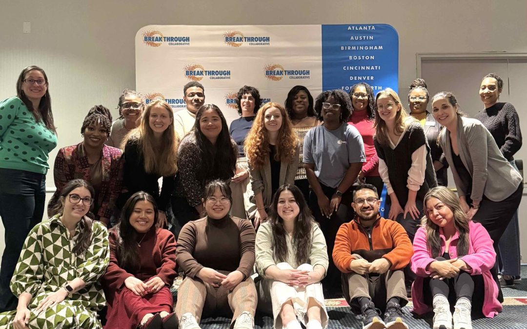 A group of 20 people of diverse backgrounds pose and smile in front of a Breakthrough banner listing several U.S. cities, indoors—celebrating educational empowerment and opportunity together. Most are seated or kneeling, with others standing behind them.