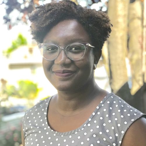 A woman wearing glasses and a gray sleeveless dress with white polka dots stands outdoors, smiling softly—her expression radiates educational empowerment against a backdrop of blurred green foliage and sunlight.