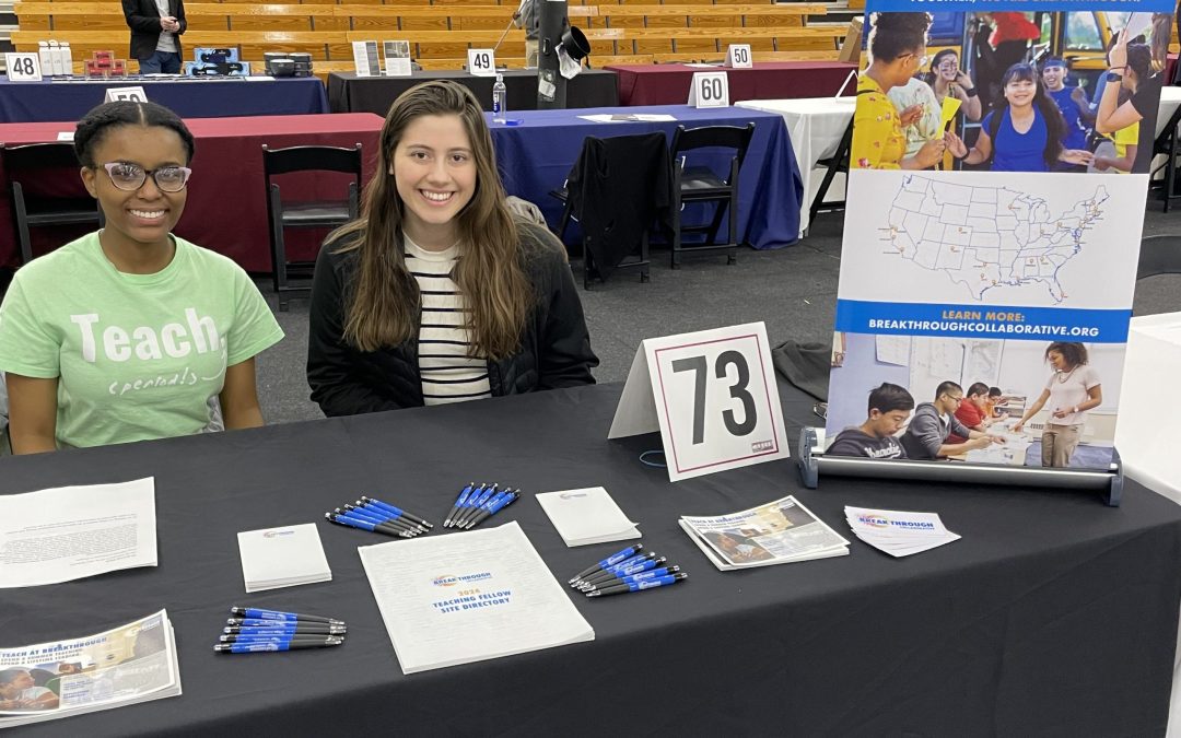 Two women sit behind a table at a Breakthrough Collaborative event booth, offering educational enrichment brochures and flyers. A large banner, the number 73 sign, bleachers, and other booths highlight this unique education opportunity.