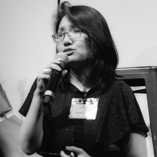 A woman with glasses, wearing a name tag, speaks into a microphone—her expression reflecting educational empowerment. She has shoulder-length hair and wears a dark, short-sleeved top. The black and white image captures this inspiring moment.