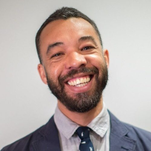 A man with a beard and mustache smiles broadly at the camera, embodying educational empowerment. He wears a blue blazer, light gray shirt, and navy tie with a subtle pattern, standing against a plain light background.