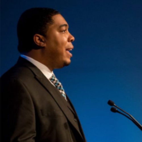 A man in a dark suit and patterned tie speaks into a microphone at a podium against a blue background, highlighting the importance of educational empowerment.