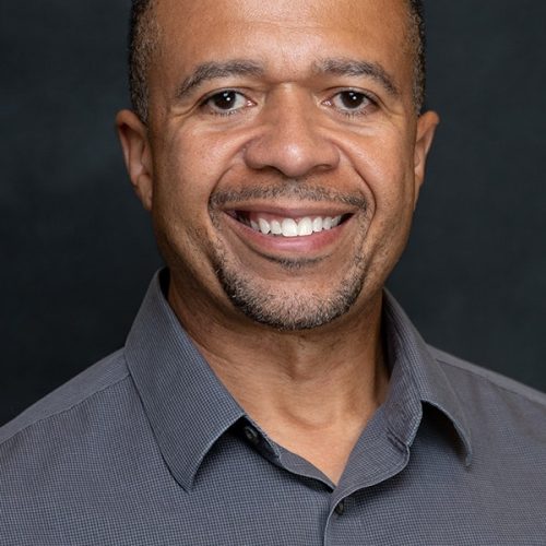 A smiling man with short hair and a goatee, wearing a gray collared shirt, poses in front of a dark background, embodying the spirit of educational enrichment and opportunity.