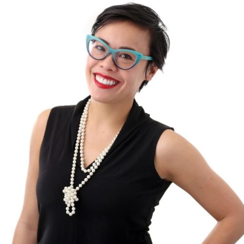 A smiling person with short black hair, wearing blue glasses, a sleeveless black top, a long pearl necklace, and red lipstick, poses against a white background, embodying the spirit of educational enrichment and opportunity.