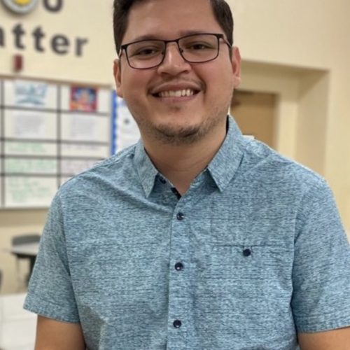 A man wearing glasses and a light blue short-sleeve button-up shirt smiles at the camera in a classroom setting, reflecting educational empowerment amid bulletin boards and tables in the background.