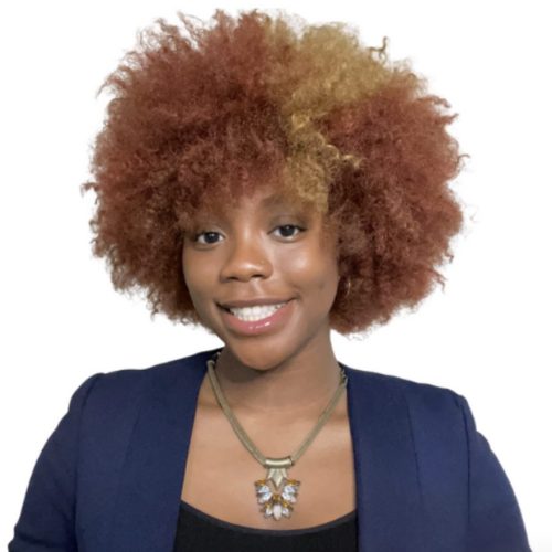 A woman with curly, reddish-brown and blonde hair smiles at the camera, embodying educational enrichment. She wears a navy blazer, black top, and a statement necklace with light-colored stones against a plain white background.
