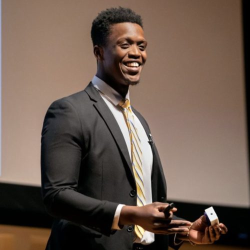 A man in a suit and striped tie smiles while giving a presentation on stage, holding a remote control in one hand and gesturing with the other as he discusses educational enrichment opportunities.