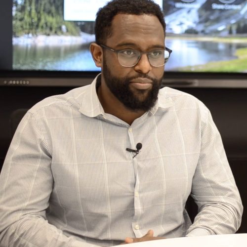 A man with short hair, a beard, and glasses sits at a table in a light-colored, checked shirt with a microphone clipped on, ready to discuss educational enrichment. A screen displaying a nature scene is visible in the background.
