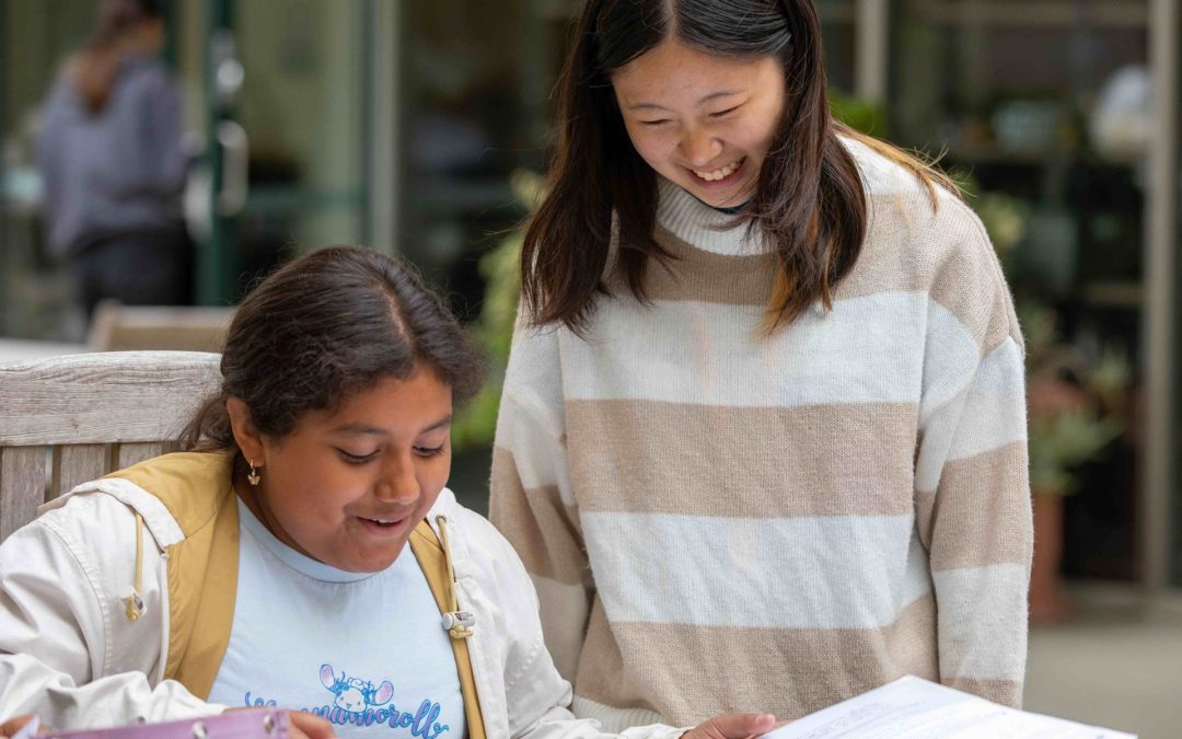 Two young women interact outdoors; one sits looking at an open binder, while the other stands beside her, smiling. They appear engaged in a friendly conversation or study session, highlighting the value of educational enrichment and shared opportunity.