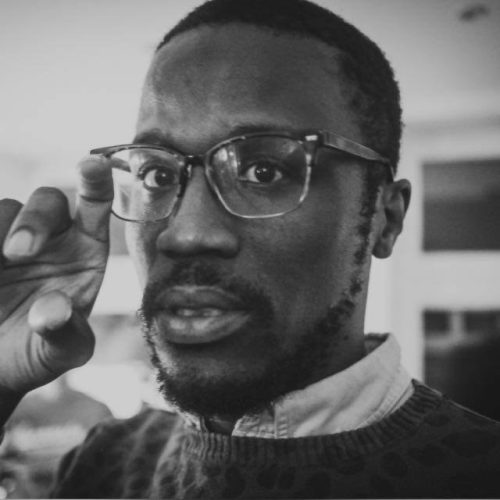 A man wearing glasses and a patterned sweater looks directly at the camera, holding his hand up near his face with his fingers slightly apart, capturing a thoughtful moment that suggests the opportunity for educational enrichment. The image is in black and white.