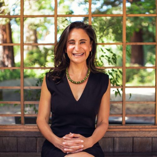 A woman with long brown hair, wearing a black dress and a green beaded necklace, sits on a wooden bench, smiling with her hands folded—radiating confidence and educational empowerment against a backdrop of wooden lattice and greenery.