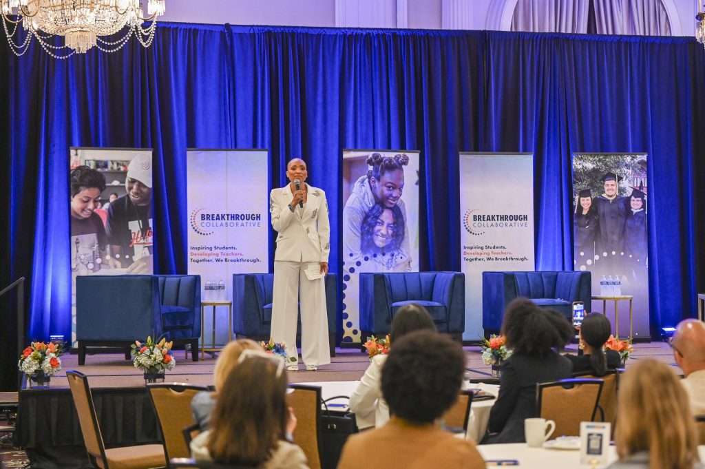 A speaker in a white suit stands on stage with a microphone at a conference. Behind her are blue curtains, banners with the “Breakthrough Collaborative” logo, and images of students, highlighting educational empowerment. Attendees are seated at tables in the foreground.