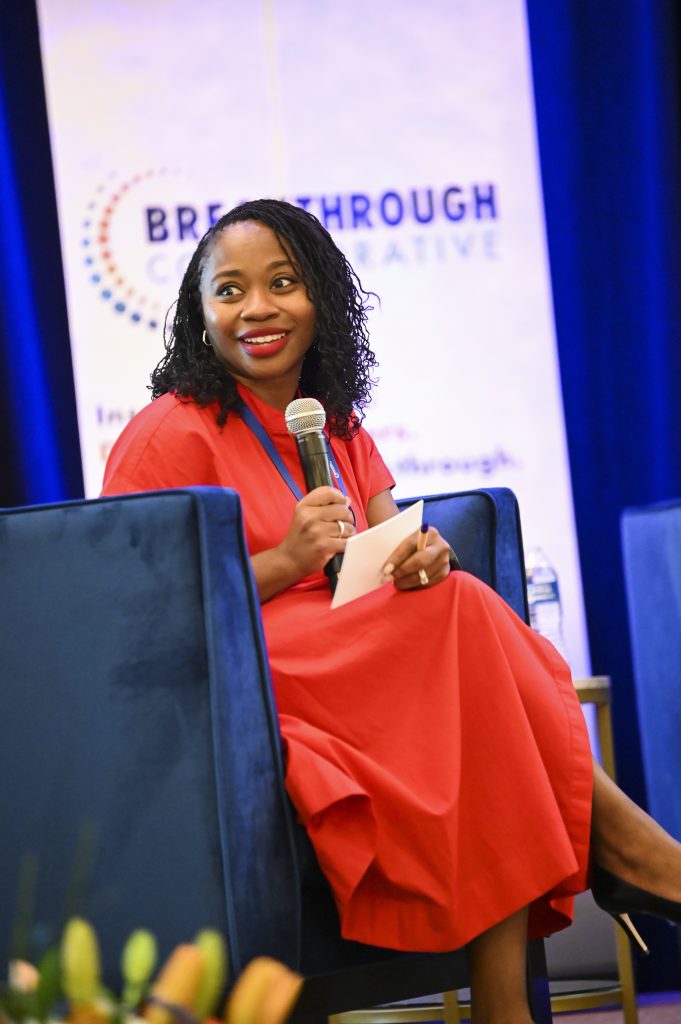 A woman in a bright red dress holds a notecard and smiles while seated on a blue chair at an educational empowerment event. A sign in the background reads Breakthrough Collaborative.