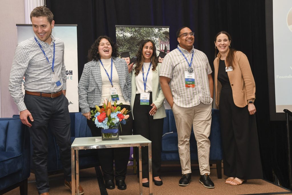Five people with name tags stand smiling and laughing on a stage next to a small table with a flower arrangement. Two blue chairs, a black curtain, and conference signage highlight the opportunity for educational empowerment.