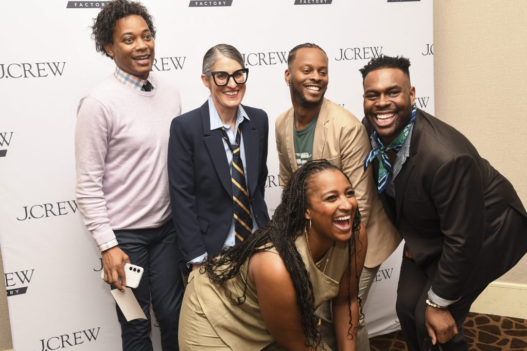 Five people smiling and posing together in front of a J.Crew and J.Crew Factory step-and-repeat backdrop at an indoor event, celebrating educational enrichment. Four are standing, while one woman crouches, laughing in the front.