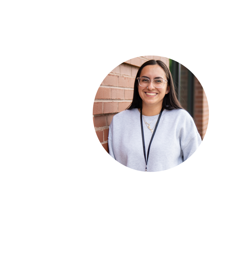 A smiling woman with long dark hair and glasses stands against a brick wall, wearing a light gray sweater and a lanyard—embodying educational empowerment. The photo is cropped in a circular frame on a white background.