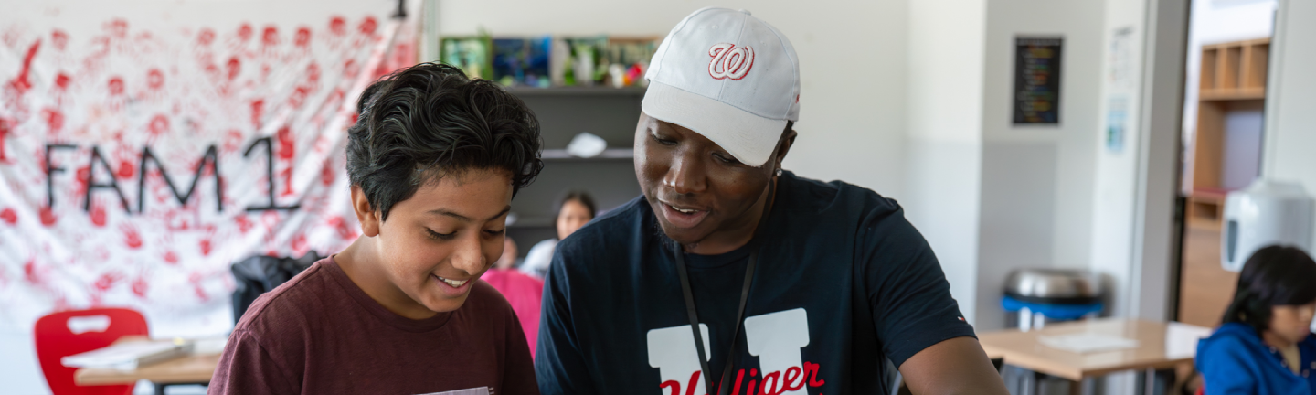 A smiling adult wearing a baseball cap and t-shirt helps a young student with dark hair in a classroom, fostering educational empowerment. Other students and colorful classroom decorations are visible in the background.