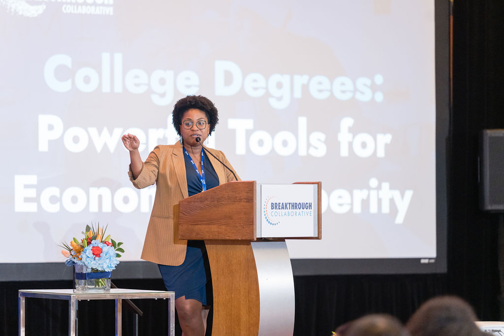 A woman speaks at a podium labeled Breakthrough Collaborative in front of a large screen displaying College Degrees: Powerful Tools for Economic Prosperity, highlighting educational empowerment. A table with a flower arrangement sits beside her.
