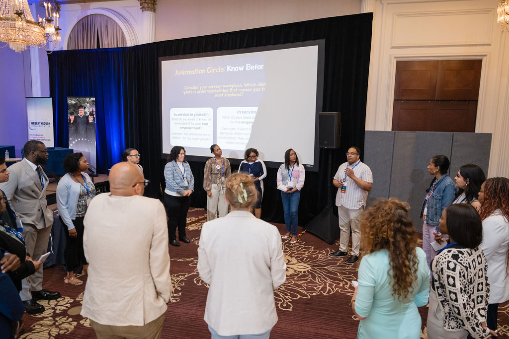 A diverse group of people stand in a circle in a conference room, listening to a speaker. Behind them, a presentation is projected onto a screen, creating an atmosphere of professional collaboration and educational empowerment.