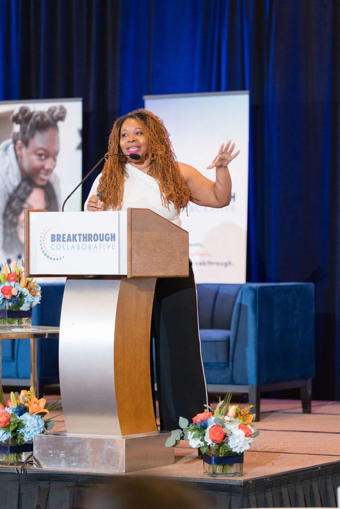 A woman with long curly hair speaks passionately at a podium labeled Breakthrough Collaborative, highlighting educational enrichment. Colorful flower arrangements and banners decorate the background as she gestures while addressing the audience.