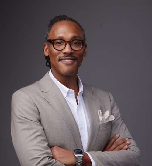 A man wearing glasses and a beige suit with a white shirt and pocket square stands against a gray background, smiling with arms crossed, embodying confidence and educational empowerment.
