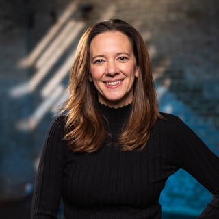 A woman with long brown hair, wearing a black ribbed sweater, smiles while standing in front of a blurred brick wall with sunlight streaming through, embodying the spirit of educational empowerment and opportunity.