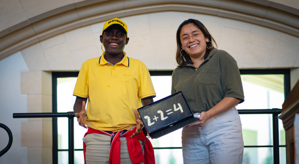 Two people smiling indoors, one in a yellow shirt and cap, the other in green, hold a small chalkboard displaying 2+2=4—celebrating educational empowerment through simple moments of learning together.