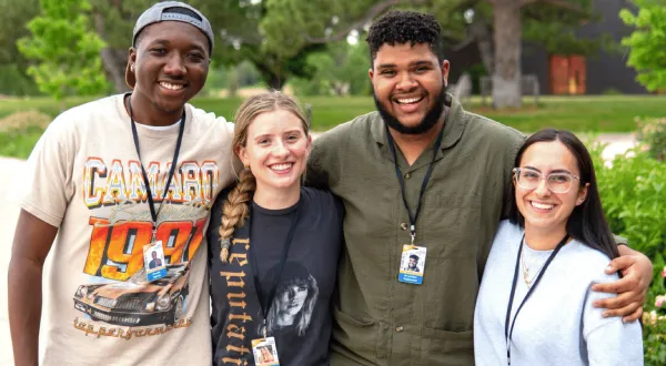 Four young adults stand outdoors, smiling with their arms around each other. Wearing ID badges and casual clothing, they enjoy an opportunity for educational enrichment amid green trees and grass in the background.