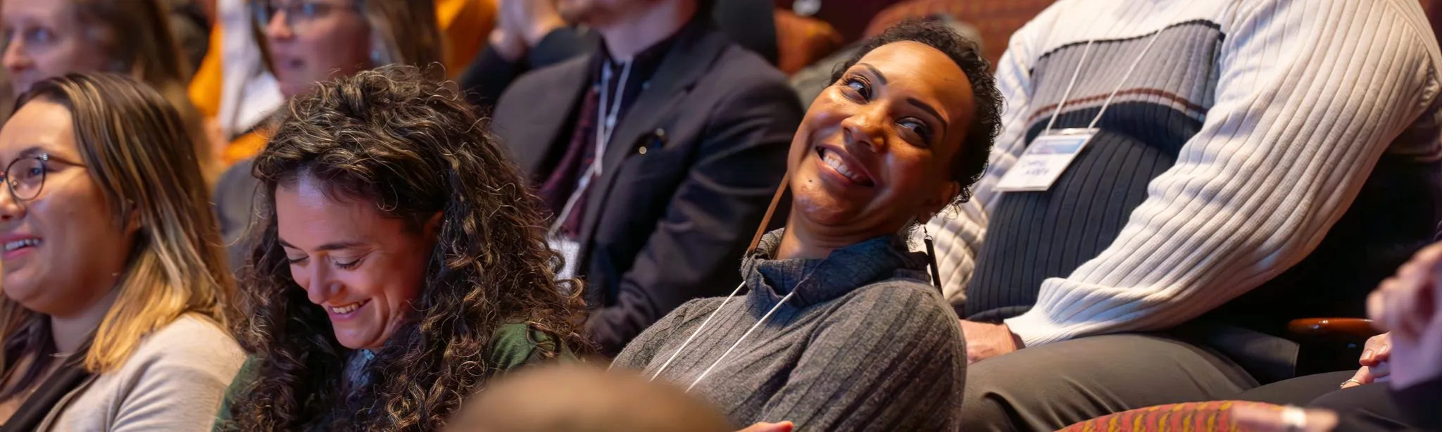 careers-hero A group of people sitting in an audience, smiling and laughing. Two women in the foreground appear particularly joyful, embracing the moment and highlighting the power of educational empowerment within this lively gathering.