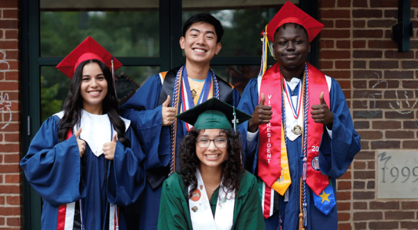 Four smiling graduates in caps and gowns pose outside a brick building, celebrating educational empowerment. Three wear blue gowns and red caps, one wears green; two give thumbs up, and all display graduation cords and sashes.