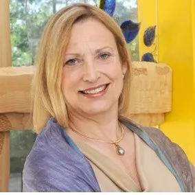 A smiling woman with shoulder-length blonde hair wearing a light gold top, a gold necklace with a pendant, and a lavender shawl, poses indoors by a wooden structure with yellow and blue accents, reflecting educational empowerment.