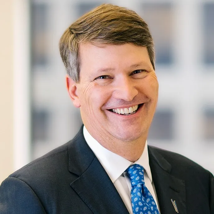 A man in a dark suit, white shirt, and blue patterned tie smiles at the camera. He has short brown hair and stands in front of a blurred indoor background, embodying educational empowerment and opportunity.