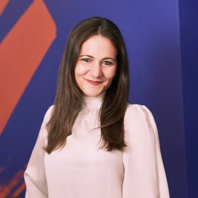A woman with long brown hair wearing a light pink blouse smiles at the camera, standing in front of a blue and orange abstract background, embodying the spirit of educational empowerment.