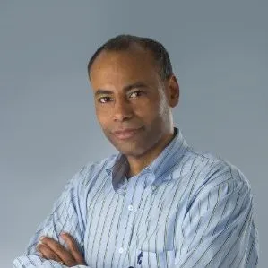 A man with short hair wearing a blue and white striped button-up shirt stands with arms crossed, embodying educational empowerment as he looks confidently at the camera against a plain gray background.