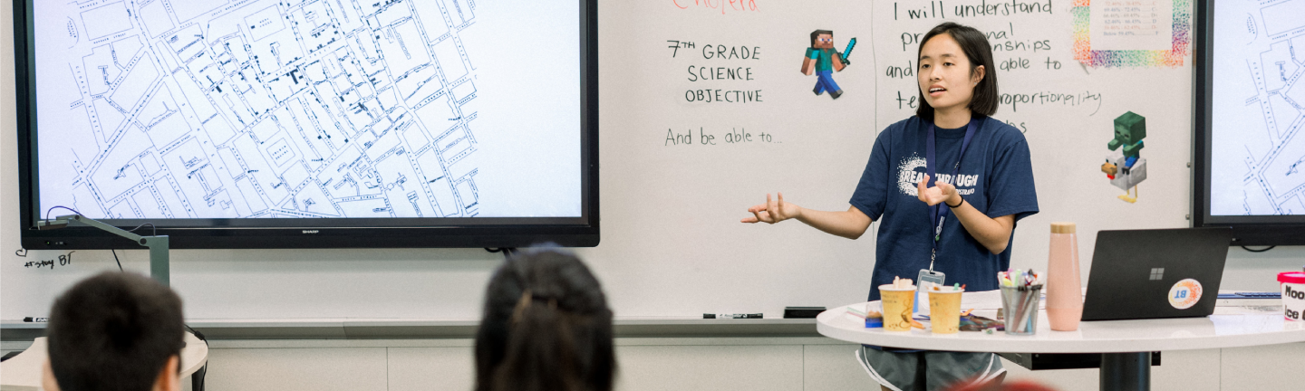 donate-hero-banner A teacher stands at the front of a classroom, gesturing while speaking to students. Behind her are two large screens—one displaying a map, the other a whiteboard with handwritten science objectives for educational enrichment.