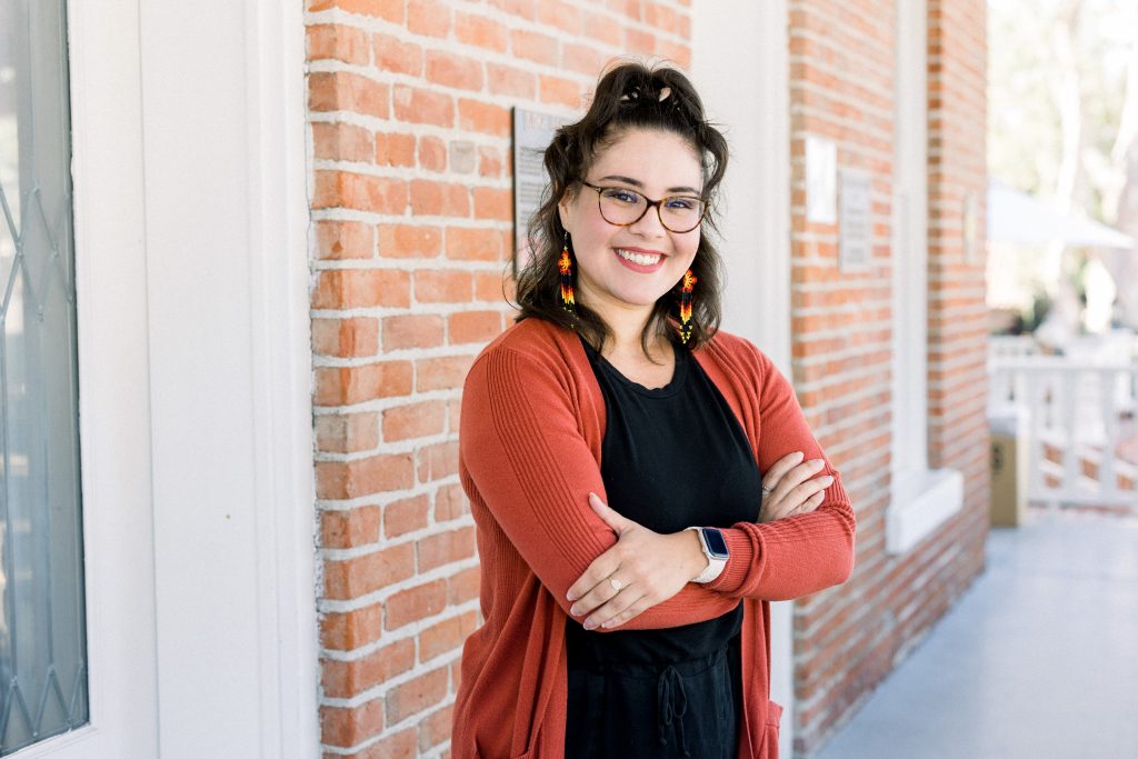 A woman with glasses, curly hair, and colorful earrings smiles with arms crossed. She wears a red cardigan over a black outfit and stands in front of a brick wall on a sunny day.