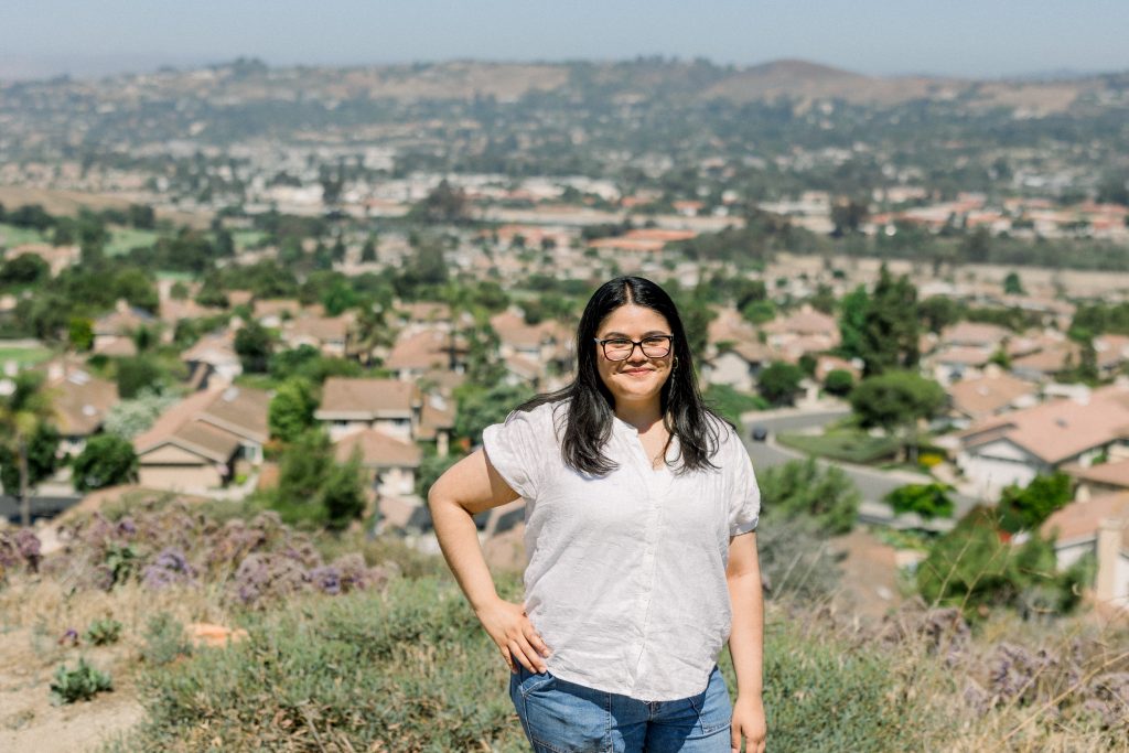 A woman with long dark hair and glasses, wearing a white shirt and jeans, stands outdoors on a hill overlooking a suburban neighborhood with houses, greenery, and distant hills under a clear sky.