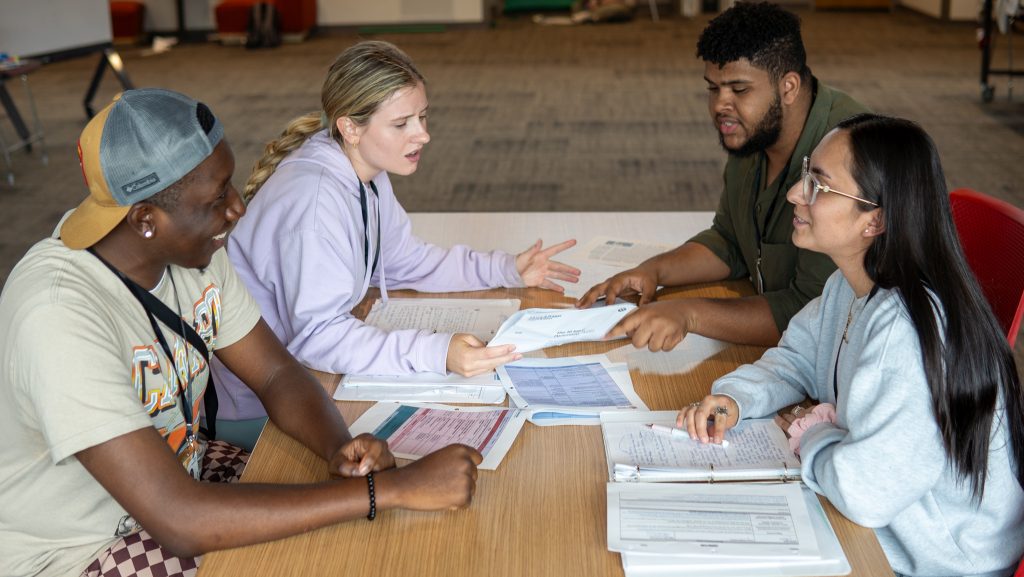 Four college students sit around a table with notebooks and papers, discussing something with engaged and animated expressions, suggesting teamwork or a group project in a classroom setting.