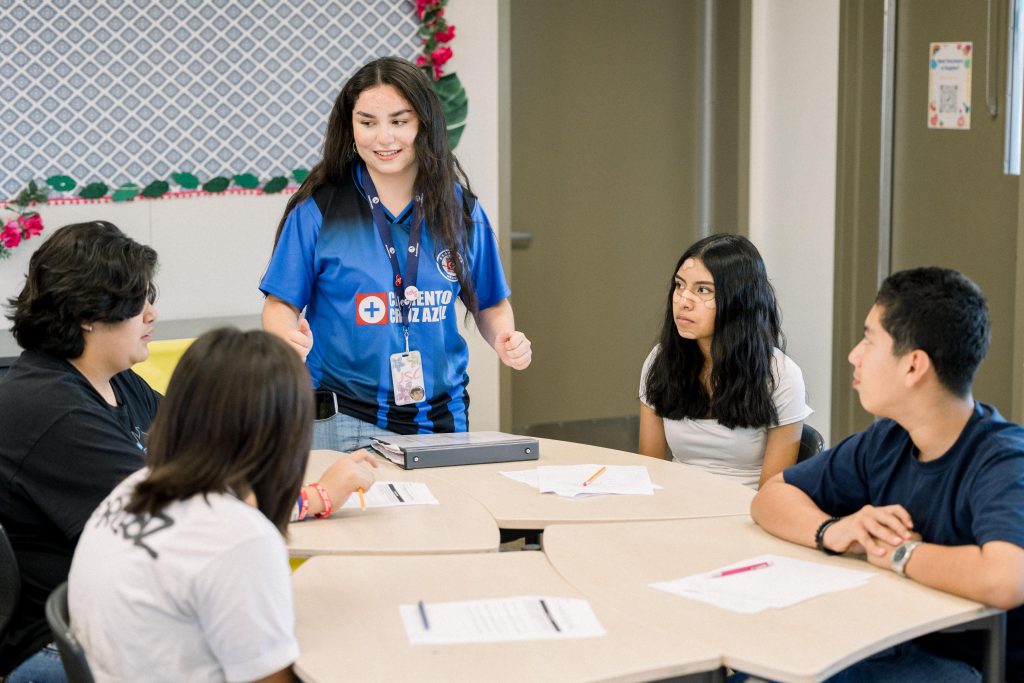 A young woman stands and speaks to four seated students around a table in a classroom. Papers and pencils are on the table, and everyone appears engaged in discussion. The room is bright and decorated with colorful accents.