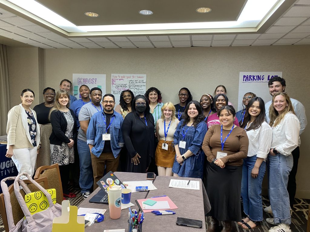 A diverse group of people, smiling and standing together in a conference room. Behind them are posters labeled Energizer and Parking Lot. A table with papers, drinks, and supplies is in the foreground.