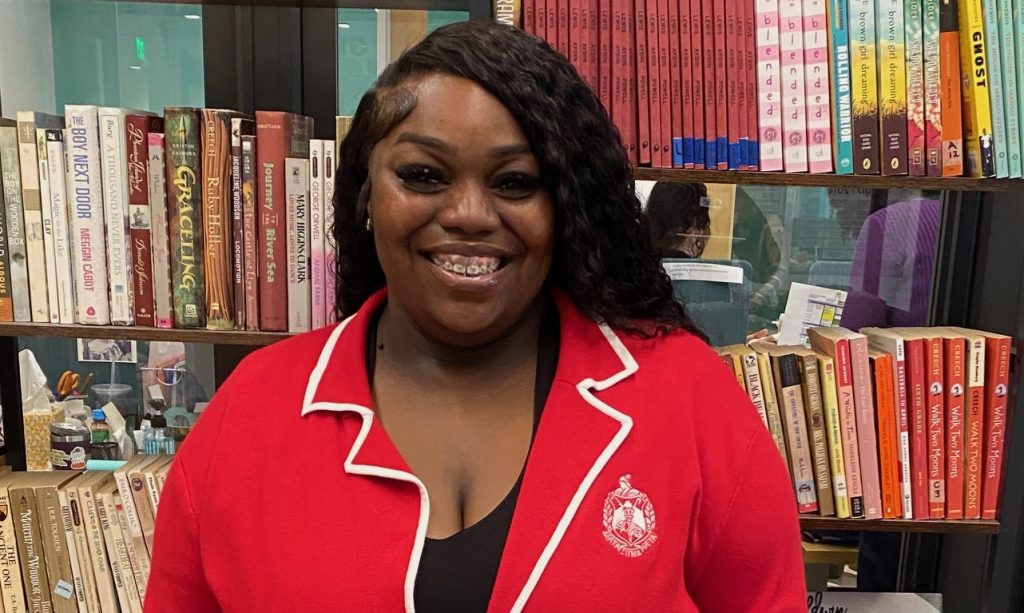 A smiling woman wearing a red jacket stands in front of a bookshelf filled with colorful books arranged vertically and horizontally.