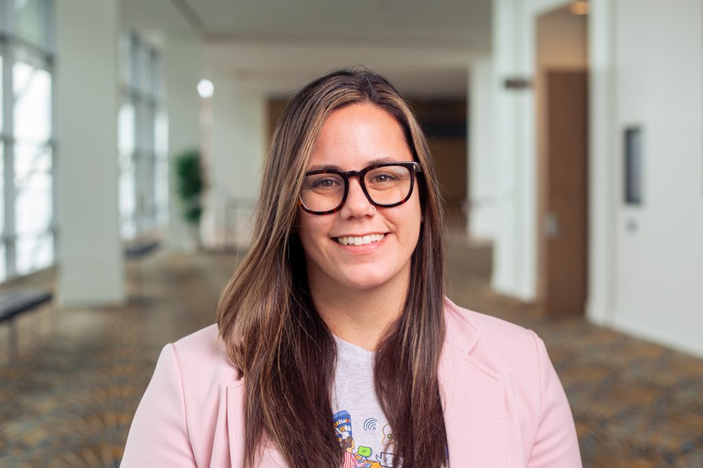 A woman with long brown hair and glasses smiles at the camera. She is wearing a light pink blazer and a t-shirt. The background shows a bright, spacious hallway with large windows and patterned carpet.