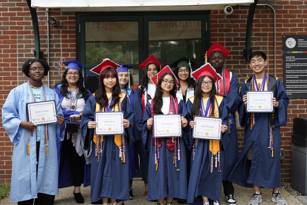 A group of diverse high school graduates in caps and gowns stand outside, smiling and holding certificates, posing for a photo in front of a brick building.